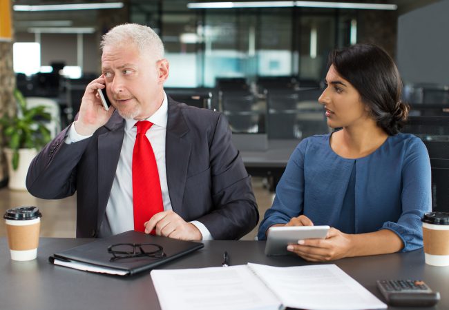 Busy boss talking on phone at meeting while his female assistant waiting for decision. Concentrated senior businessman using mobile phone to connect partner. Work with prominent customers concept