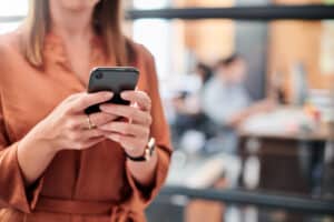 A person wearing a brown shirt uses a smartphone, with an office environment slightly blurred in the background.