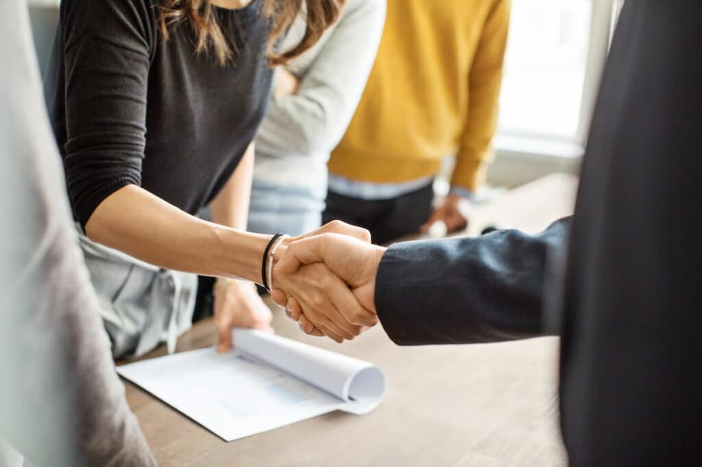 Close-up of two people shaking hands across a meeting table, with documents and colleagues visible in the background, representing a business agreement or professional partnership.