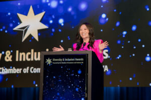 A woman in a pink outfit is speaking at a podium during the Diversity & Inclusion Awards event, with a blue and starry background behind her.