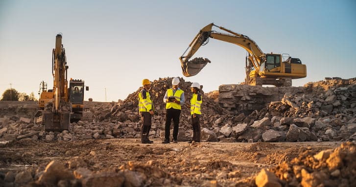 A group of mining construction workers in high-visibility vests and hard hats stand on a rocky construction mine site, with excavators in the background under a clear sky.