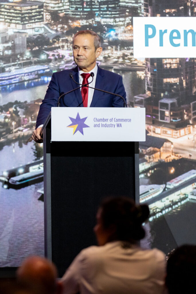 WA Premier Roger Cook is in a blue suit and red tie speaks at a podium labeled "Chamber of Commerce and Industry WA" with a cityscape background.