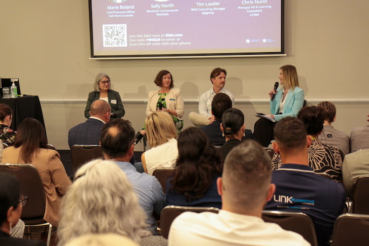 Four people seated in a row with microphones on a stage, representing a panel discussion at a well-attended event.