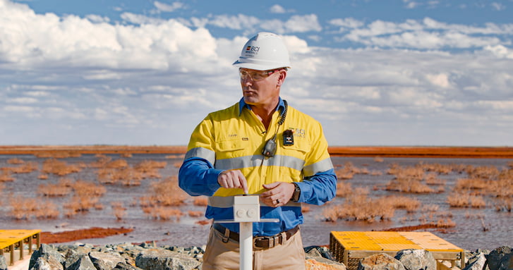 A man wearing a yellow shirt and hard hat stands in front of a vast field, surveying the landscape.