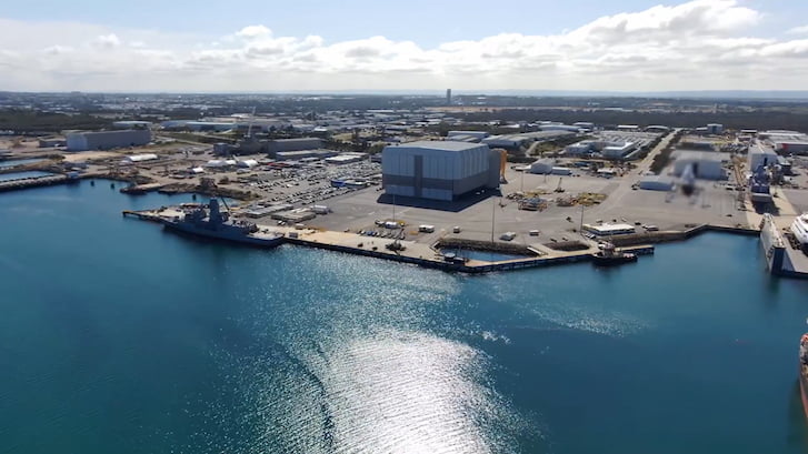 Aerial view of a harbor with a large defense ship docked, surrounded by industrial buildings and warehouses, under a partly cloudy sky.