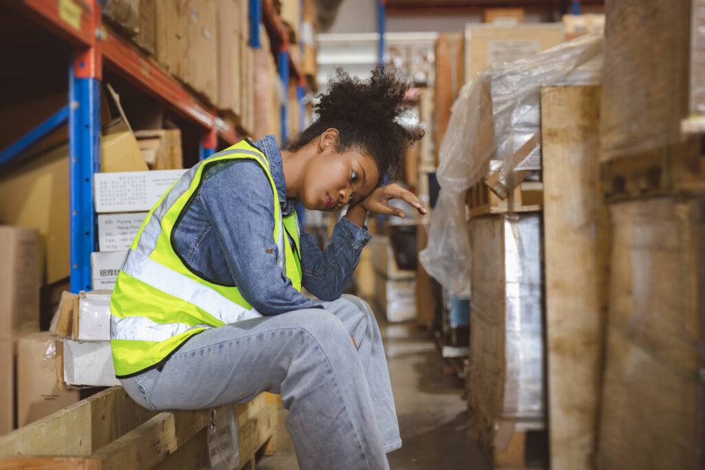 A woman seated on a box inside a warehouse, surrounded by shelves and various storage items.