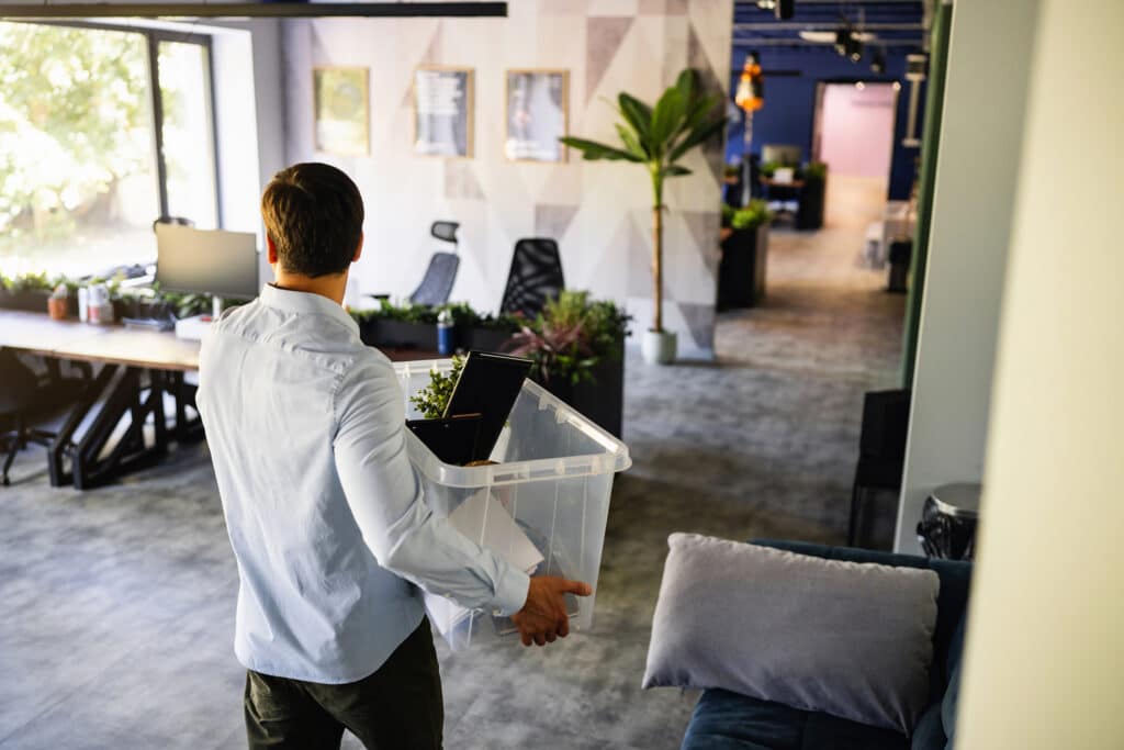 Employee carrying a box of belongings through an office, representing workplace redundancy, business restructure and organisational change.
