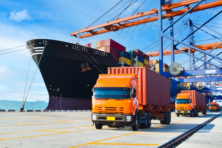 Trucks parked in front of a large container ship at a port, symbolising the vital role of trade in global logistics.