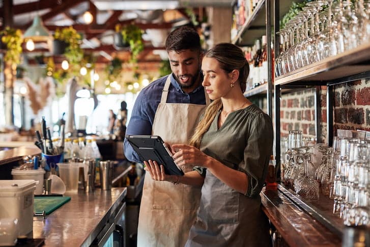 A man and woman stand at a bar, engaging with a tablet, representing a small business environment.