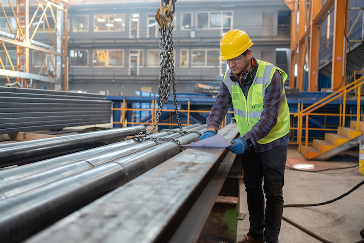 A man in a hard hat and safety vest inspects steel pipes at a manufacturing facility.