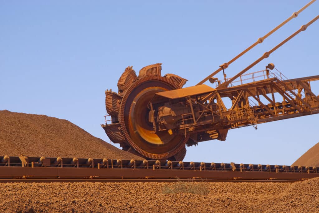 A large machine lifts a substantial pile of iron ore at a mining site.