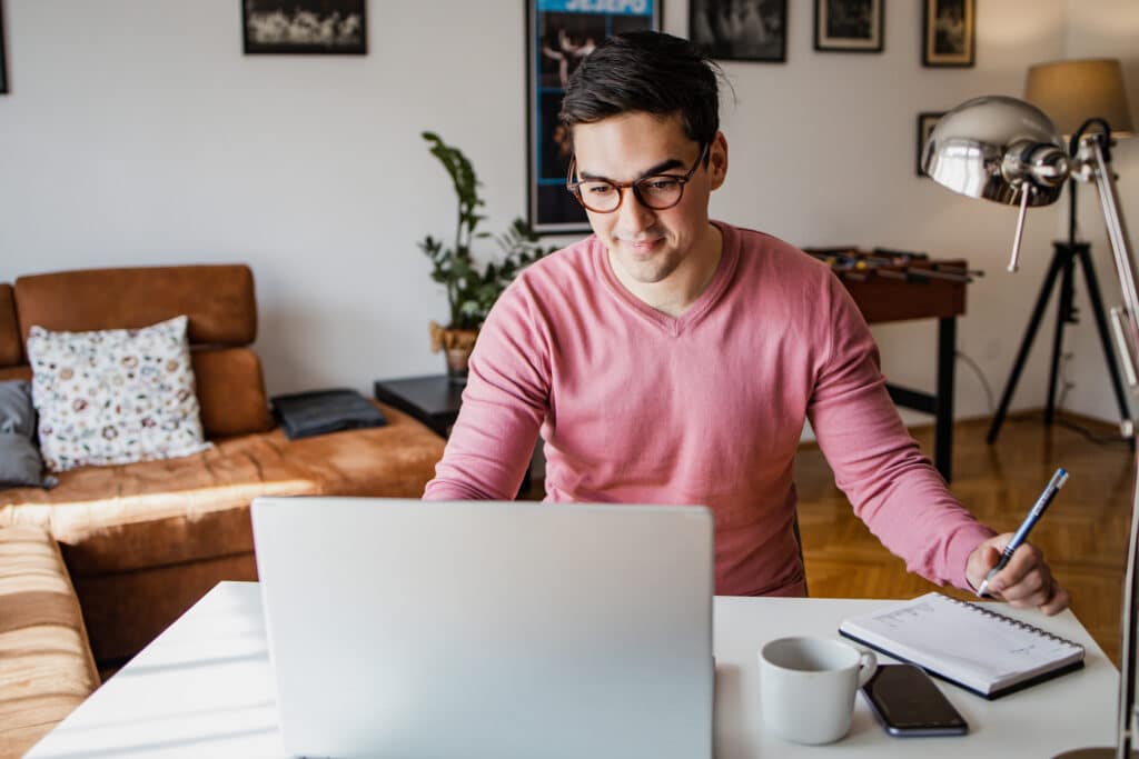 Young professional working remotely from home, wearing glasses and a pink sweater, taking notes during a video call on a laptop at a white desk with coffee, smartphone, and notebook.
