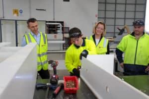 Apprentice using a power tool to assemble cabinetry while supervisors in high-visibility vests observe at a manufacturing workshop.