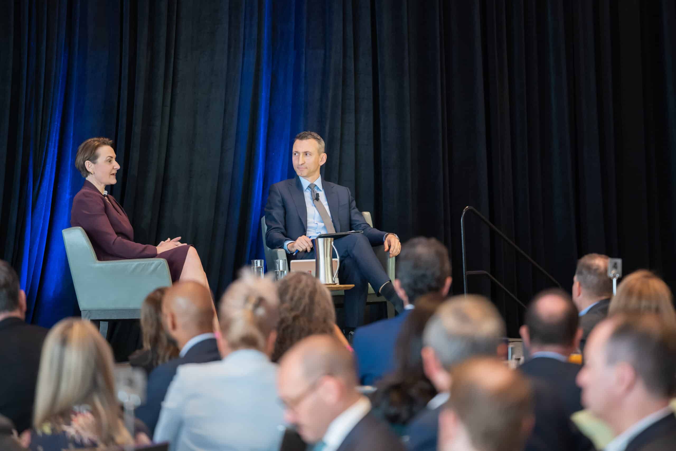 Two individuals seated on chairs facing a large audience in a public speaking setting.