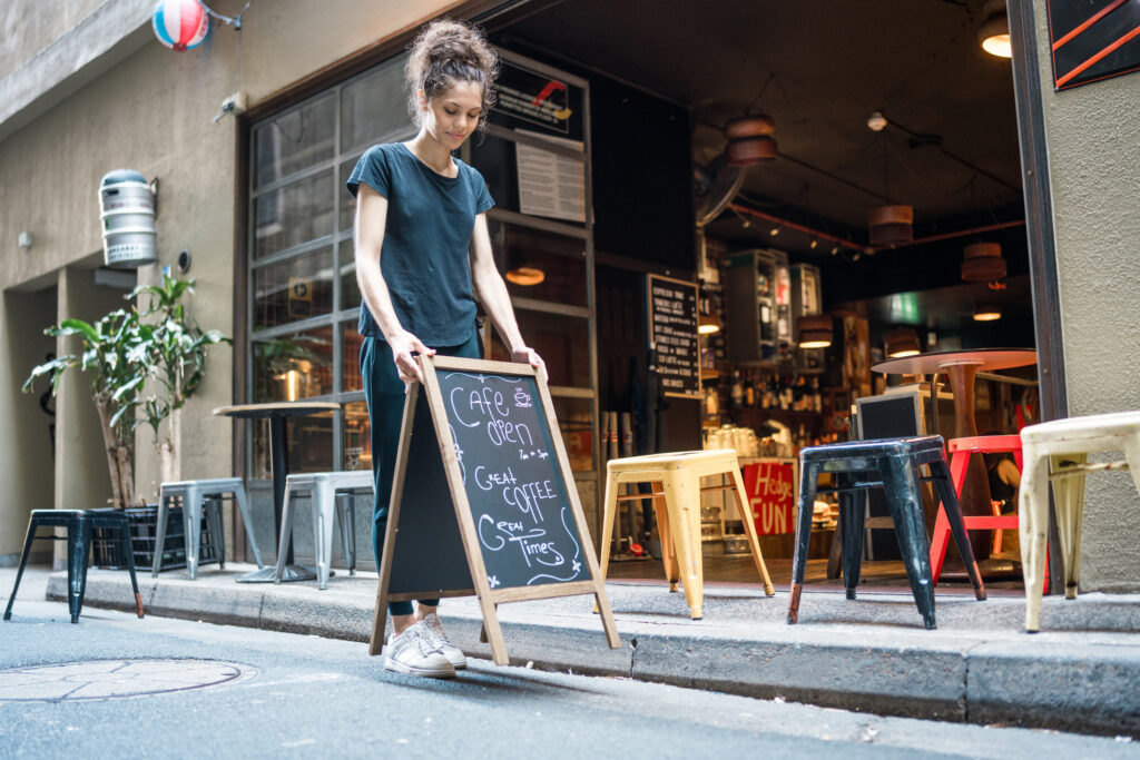 A woman stands in front of a small business restaurant, next to a chalkboard displaying the daily specials.