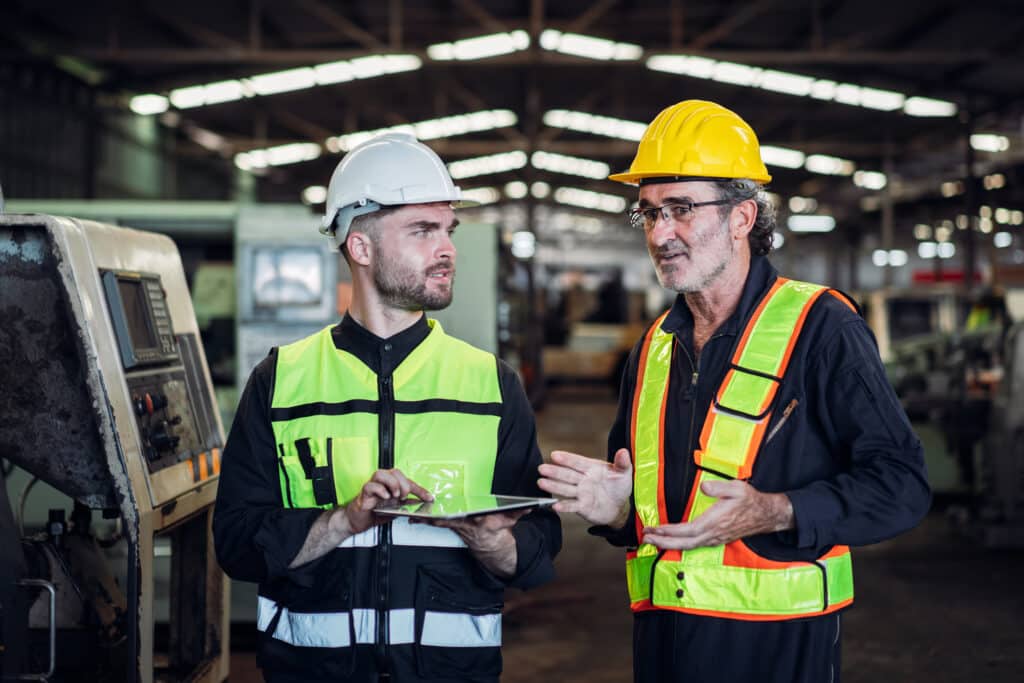 Two men in safety vests engaged in conversation inside a factory setting.