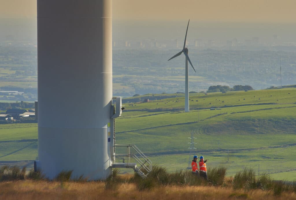 A group of people stands beside a wind turbine, discussing project development and renewable energy initiatives.
