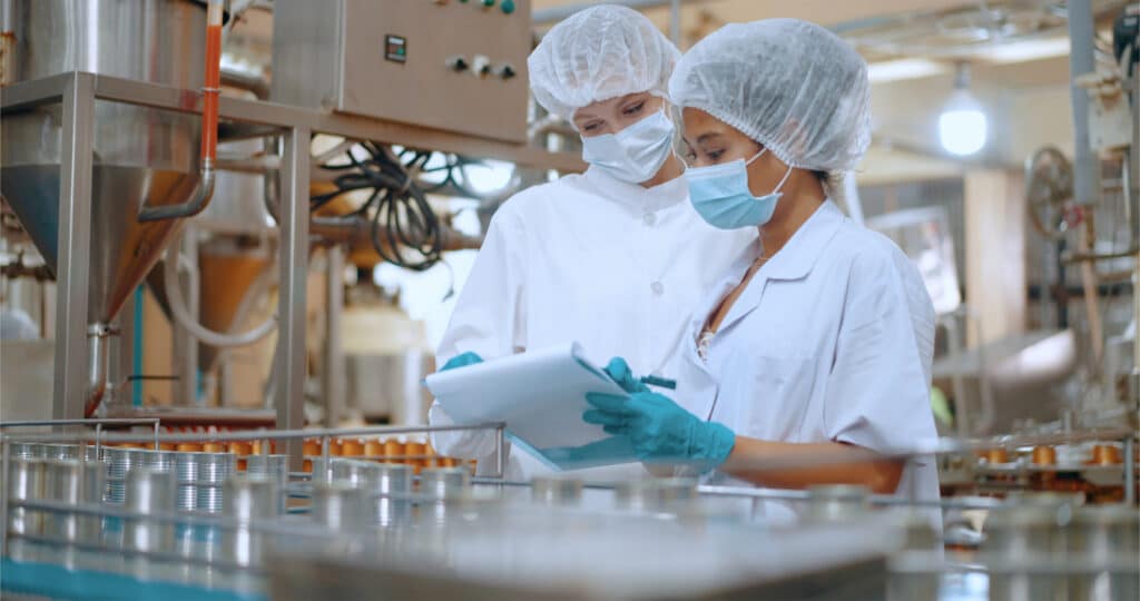 Two professionals in white lab coats collaborate on a tablet in a beverage manufacturing setting.