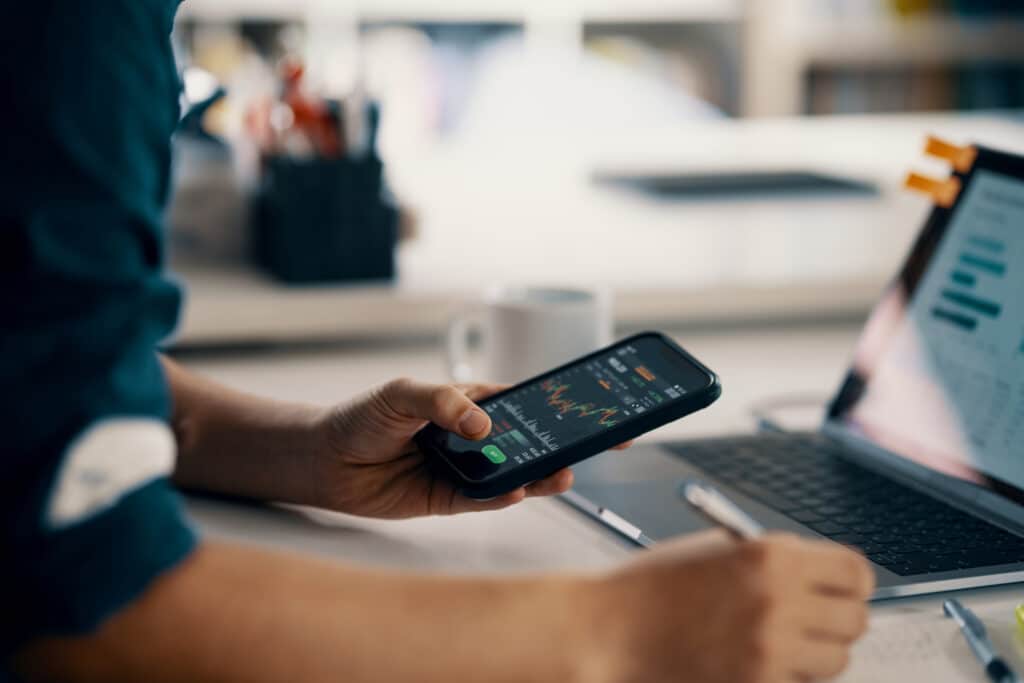 Man using a smartphone at a desk with a laptop, focused on investment readiness tasks.