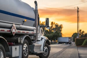 Industrial carrier white day cab big rig semi truck tractor transporting flammable fuel liquid in tank semi trailer running on the local road at evening sunset time.