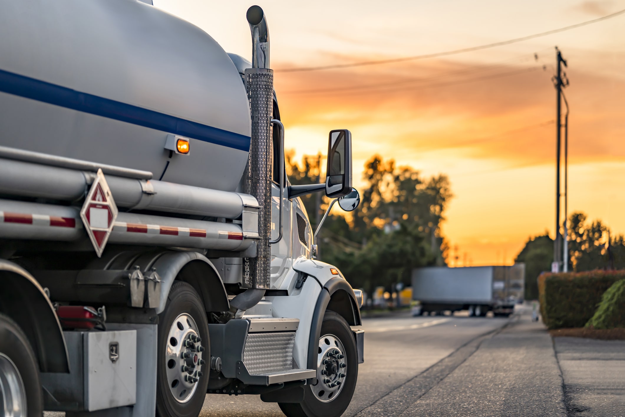Industrial carrier white day cab big rig semi truck tractor transporting flammable fuel liquid in tank semi trailer running on the local road at evening sunset time.