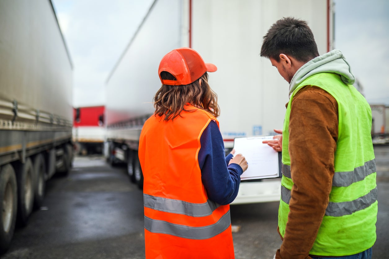 Two logistics workers in high-visibility vests reviewing paperwork beside freight trucks, representing transport operations, supply chain coordination and freight management.