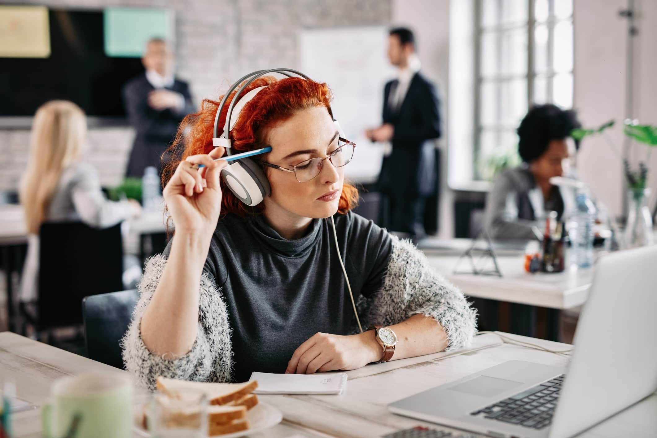Employee wearing headphones and working on a laptop in a busy office environment, highlighting workplace training, professional development and focused learning.