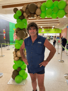 Tony Galati stands beside a green and gold balloon arch in a shopping centre, with the Spudshed store visible in the background.