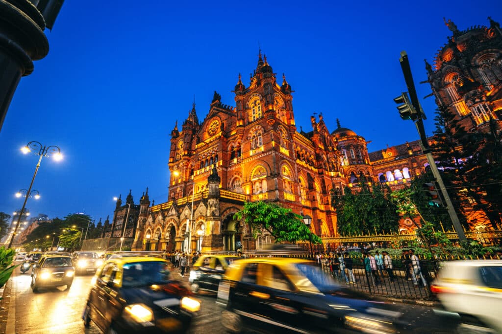 Chhatrapati Shivaji Maharaj Terminus illuminated at night in Mumbai, India, with taxis and traffic passing outside.