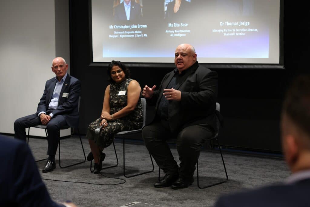 Three panellists sit in front of a screen listing speakers, while the man on the right speaks and gestures to the audience.
