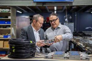 Two men in a workshop examine a metal component they are holding, with coiled black cable and similar parts on the bench. The man on the left is WA Premier Roger Cook