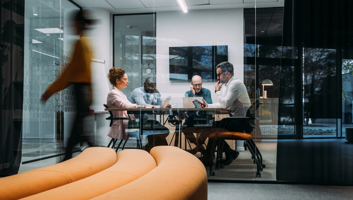Four colleagues meet around a table in a glass-walled office as another person walks past.